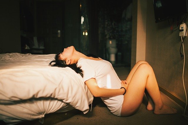 Žena v bielom tričku opretá o posteľ so zaklonenou hlavou 